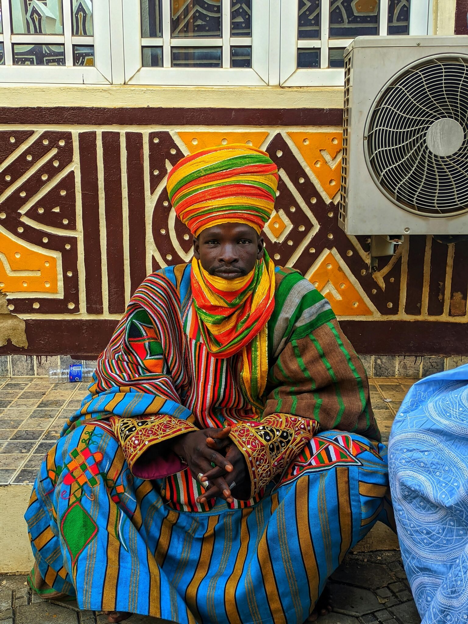 A man in vibrant traditional attire sitting in front of tribal wall art, Nigeria.